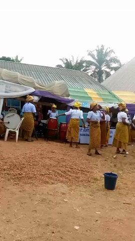 The video captures a group of women dressed in matching outfits performing a dance outdoors. They wear white tops, skirts with an intricate pattern, and matching headwraps. Some of the women are playing drums as accompaniment for the dancers.

The setting appears to be a gathering or event, with tents set up in the background. A table with the words "ST. ANTHONY OF PADUA STATION" is visible behind the dancers. The ground is covered with small objects, possibly pebbles or nuts. A dark-colored bucket sits in the foreground. Trees are visible in the distance, adding to the outdoor scenery.