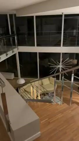 The video shows a wide shot of a modern interior design from an elevated perspective. A wooden staircase with stainless steel handrails leads down to a living area. The living area is furnished with a light-colored sofa, a glass-topped table, and a spherical lamp. Large windows are visible in the background, showing darkness outside. The design includes a second-story level with glass railings. The general ambiance appears to be quiet and dimly lit.