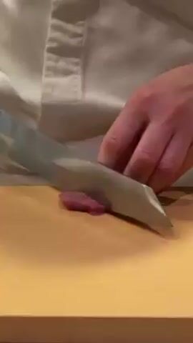 A medium shot depicts a person in a white shirt slicing a piece of red meat on a wooden cutting board. The person uses a large, sharp, silver knife, carefully guiding it to make thin cuts. The hand not holding the knife is positioned to stabilize the meat. The background is plain and out of focus, drawing attention to the food preparation process.