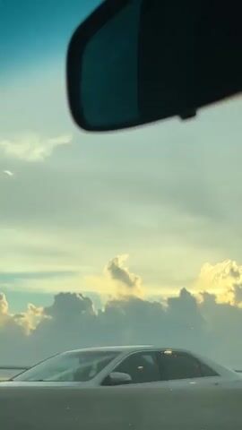 The video shows a view from inside a vehicle looking out the windshield. The sky is visible with a mixture of clouds. Sunlight is breaking through the clouds, creating a hazy effect. In the distance, a bridge or overpass can be seen with structures resembling traffic lights or poles along its length. The bottom of the rearview mirror of the vehicle is visible in the upper portion of the frame.