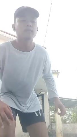A person wearing a light blue long-sleeved shirt, black shorts, and a black baseball cap stands outside next to a tan building. The person is in the process of performing a martial arts move, raising their right leg in a knee strike motion while keeping their left leg on the ground. Their arms are positioned defensively in front of their body. The building has a white overhang supported by a yellow pillar, and a black metal gate is visible behind the person. The sky is overcast.