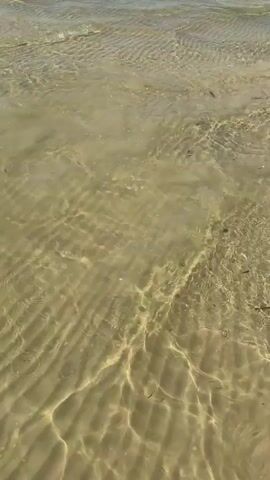 The video shows a shallow, sandy lakebed with clear water rippling over it. The camera pans to reveal a sunny day with a blue sky and scattered clouds. In the distance, on the water, are several inflatable water toys, including a large yellow banana boat and a blue and white inflatable, possibly a jet ski accessory. Several people are visible on these inflatables, enjoying the water. Further in the background, a water slide complex is visible along the shoreline, with more people scattered around the beach and in the water. The scene depicts a recreational area at a lake on a summer day.