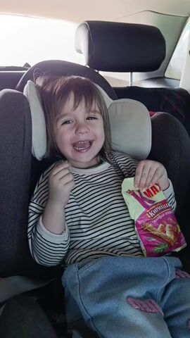 A young child is seated in a car seat inside a vehicle. The child is wearing a striped shirt and jeans and is holding a package of snack crackers. The child is smiling broadly, revealing missing teeth. The car seat is gray, with white headrests. The interior of the car is visible, including the back of the front seat. A song is playing in the background.