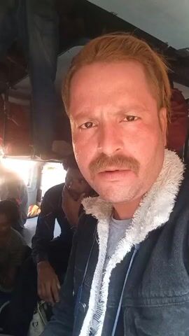 The video features a man with a mustache and reddish-blonde hair, wearing a denim jacket with a faux fur collar and a gray t-shirt. He is looking directly at the camera. The background shows other people sitting, including a child in a striped shirt and a man with his hand on his face. Overhead, there is luggage and a person standing. The scene appears to be inside a crowded vehicle, possibly a train. There is a distinct sound of train wheels on the tracks.
