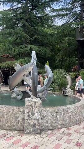 This video showcases a whimsical outdoor fountain featuring large, realistic sculptures of fish. The central sculpture depicts several fish in dynamic poses, seemingly leaping and interacting with water jets that spray from their mouths and bodies. The largest fish is a silver-colored salmon or trout, its mouth open as water streams upwards. Other fish, including a smaller, darker fish, are integrated into the composition. The fountain is set within a circular pool with a stone-like border.

The surrounding environment is lush and green, with tall evergreen trees providing a natural canopy. In the background, a wooden structure, possibly a treehouse or elevated platform with stairs, is visible. The ground around the fountain is paved with a checkerboard pattern of red and beige tiles.

There are a few people in the scene, appearing to be visitors or patrons. One person wearing a light green t-shirt is standing near the fountain, observing it. Another person, also in a light green t-shirt, is visible closer to the background, near another smaller fish fountain. A third person is partially visible to the left.

The lighting suggests it is daytime, with overcast or diffused light, implying a mild or potentially humid climate given the greenery. The overall atmosphere seems relaxed and pleasant, a recreational outdoor space. There is no visible text in the video. The scene is set in Bzypta, Abkhazia.