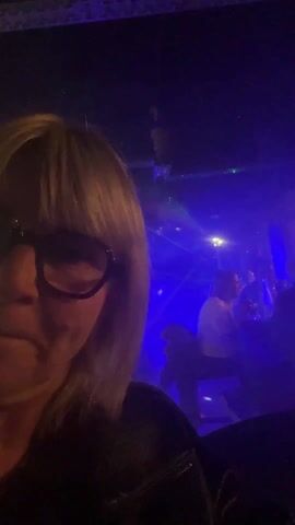 The video shows a woman with blond hair and black glasses taking a selfie in what appears to be a dimly lit bar or club. Several people are visible in the background, some standing at the bar. There are bright, colored laser lights, primarily green, red, and blue, flashing across the scene. The lighting is low, creating a club-like atmosphere. The focus remains on the woman as she seems to be enjoying the music and the environment.