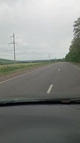 A video taken from inside a vehicle shows a straight asphalt road stretching into the distance under a cloudy sky. To the left of the road, there is a grassy field and further beyond, rolling hills. Power lines are visible on tall poles on the left side of the road. To the right of the road, there is a dense line of trees. The view is from the dashboard of the vehicle.