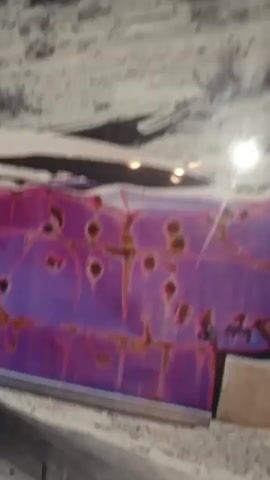 The video shows a close-up of a damaged, rusty, purple car parked in a desert environment. The car appears to be an older model, possibly a sedan, with several bullet holes visible on its side. The paint is peeling and rusted, revealing the metal underneath. The background is a barren, rocky landscape with sparse vegetation, suggesting a dry and arid climate. The lighting in the video is harsh and bright, casting strong shadows. The camera slowly pans across the car, focusing on the bullet holes and the overall state of decay. There are no people visible in the video.