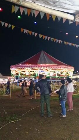 Here is a description of the video:
The video shows a carousel ride at night. The carousel has a red and white striped roof, with white lights outlining the edges. There are people riding on the carousel and people standing around it. The ground is dirt and grass. There are strings of pennant flags and colored lights overhead. The scene is set up as a fair or festival. The video is shaky, and the perspective is from under an awning of some kind. The loud music suggests a festive environment.