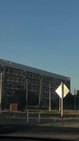 The video is a first-person perspective of driving through a somewhat rural area. The scene is dominated by a large, weathered apartment building on the left side of the frame, followed by a dirt road with sparse vegetation and utility poles. As the car moves forward, the view transitions to a street lined with smaller houses and more utility poles. The sky is clear and blue. A road sign with a diamond shape is visible in the foreground at the beginning of the video, followed by a sign with a triangle and circle. The video ends with a view of the road stretching ahead, with streetlights visible on the right side. The overall impression is one of a quiet, possibly aged, residential area during daylight hours.