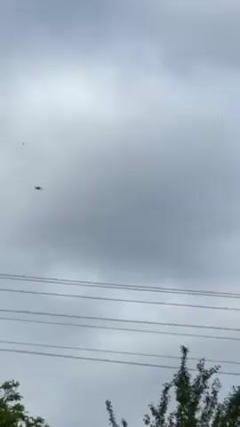 Here is a detailed description of the video:

The video captures a distant view of a fighter jet soaring across a cloudy sky. The aircraft appears as a dark silhouette against the overcast backdrop, positioned above a series of telephone wires and partially obscured by the foliage of trees at the bottom of the frame. The surrounding trees are lush green and sway gently, indicating a slight breeze. The overall scene is somewhat muted due to the diffuse lighting typical of a cloudy day.