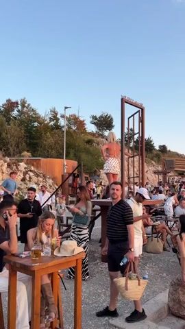 The video shows a crowded outdoor gathering at what appears to be a restaurant or bar on a hillside. A woman with blonde hair in a white polka dot dress is standing on a raised platform to the left. People are seated at tables and on benches, many holding drinks. In the background, there are trees and a structure on top of the hill. The lighting suggests it is late afternoon or early evening. The camera pans across the scene, capturing the various groups of people and the overall atmosphere of the venue. Some individuals are engaged in conversation, while others are observing the surroundings. The ground is covered in gravel and stone.
