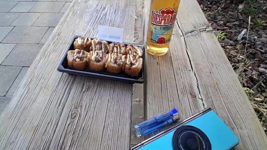 The video begins with a close-up shot of a plastic container of sushi and a bottle of Desperados beer on a weathered wooden table. A lighter and a blue object are also visible on the table. The camera then pans to the left, revealing a paved area with square tiles. The scene transitions to an outdoor environment with a grassy area, a park bench, and trees. In the background, there are buildings, a playground, and a street. The sunlight casts long shadows across the pavement, indicating it is either early morning or late afternoon. No people are visible in the video.