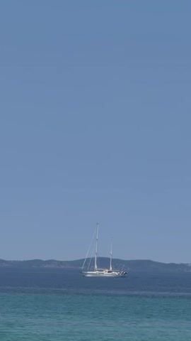 This vertical video shows a serene ocean scene with a large white sailboat as the central focus. The sailboat, with two masts and its sails furled, is positioned in the middle of the frame, gently floating on the deep blue water. In the background, a low, rolling coastline with a faint outline of hills stretches across the horizon. The sea in the foreground is a lighter, turquoise color, suggesting shallow water or a different current, and exhibits gentle ripples. The sky above is a vast expanse of clear, light blue, indicating fair weather and a bright day. The overall impression is one of tranquility and peacefulness, with the sailboat appearing to be at anchor or moving slowly. The lighting suggests it is daytime, likely midday or early afternoon given the bright, clear sky. There are no people clearly visible on the sailboat. The location appears to be a coastal area, consistent with the provided location context of Sithonia, Greece. No text is visible in the media file.