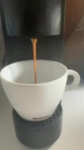 A Nespresso machine is brewing a cup of coffee. The machine is black, and the coffee is dispensed directly into a white Nespresso cup sitting on the drip tray. The coffee stream is a smooth, golden brown. The background is blurred, showing a hint of decorative items on a shelf or table.