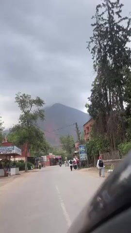 The video shows a view from a moving vehicle traveling along a paved road in a mountainous, rural or semi-rural area. The sky is overcast with heavy clouds, some of which partially obscure the peaks of the large mountain in the background. This mountain has visible reddish-brown sections, possibly indicating exposed earth or rock.

Along both sides of the road are various trees, some with dense foliage and others with more sparse branches. Scattered small buildings and shops line the road, including one on the left with a red facade and another with a green canopy. Several people, including some carrying backpacks, are seen walking along the right side of the road, heading away from the camera. Another person is walking on the left side. A motorcycle is briefly visible on the road ahead. Overhead, power lines crisscross the scene, supported by wooden poles.

Visible text on a red sign on the left side of the road reads "جلسات عائلية يومية" (Daily family sessions), suggesting a restaurant or cafe. On the right, a blue sign displays an image of a waterfall and some text, likely advertising a local natural attraction, with the word "عين" (Ain), possibly referring to a spring or a place name. The scene appears calm and everyday.