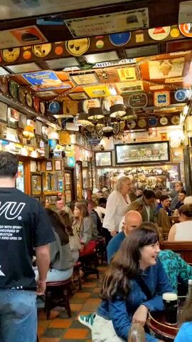 The video shows the interior of a crowded pub, likely in Ireland, given the prevalent Guinness signage. The walls and ceiling are densely decorated with various memorabilia, including signs, posters, and framed pictures. People are seated at tables and along a bar, engaged in conversation and drinking. A man in a black t-shirt with "UVU" printed on it is walking away from the camera. Several women are seated at tables in the foreground, some facing the camera. The lighting is warm and ambient, typical of a pub setting. The floor has a checkered pattern. The atmosphere appears lively and social.