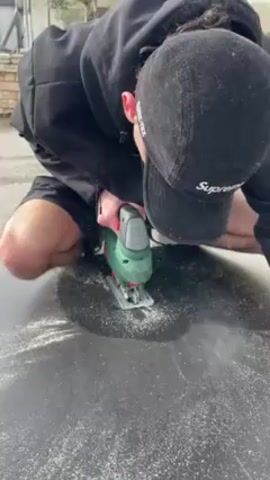 A person wearing a black hoodie, black shorts, and a black baseball cap is kneeling on a concrete surface, using a green and gray jigsaw to cut a circular hole in a black material. White dust or shavings are scattered around the cutting area. The scene appears to be outdoors, possibly in a driveway or patio area.
