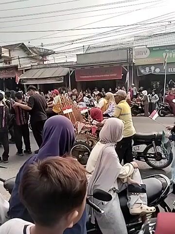 A crowd of people are gathered on a street in front of shops. A large white bus with the logo "SINAR JAYA" is visible on the left side of the frame. In the center of the scene, there is a cart loaded with what appear to be musical instruments, possibly gamelan instruments, and some red and white objects, possibly flags or decorations. Several people are standing around the cart, and others are observing from the roadside. Some individuals are on motorcycles. The environment appears to be an urban or semi-urban setting with buildings and overhead wires.