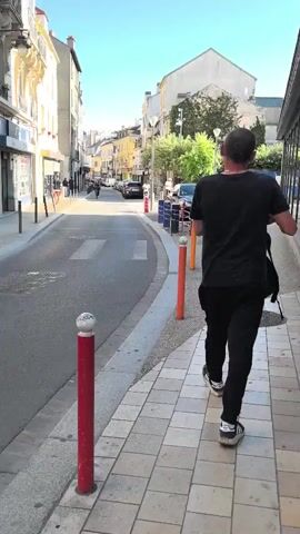 A person in a black t-shirt and black pants walks down a sidewalk on the right side of the frame. The person is walking away from the camera and toward a street. The street has a crosswalk in the middle. There are buildings on the left side of the street. The sky is blue and there are some trees on the right side of the street.
