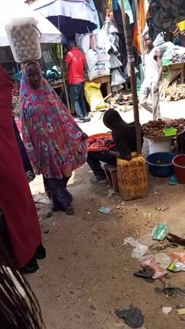 The video shows a bustling market scene. A woman wearing a floral head covering and matching robe walks past, carrying a container of eggs on her head. A young person sits on a yellow plastic container, sorting through red peppers. Other vendors are visible, selling various goods. The ground is littered with trash and debris. People walk through the market, some carrying items. A motorcycle passes in the background.