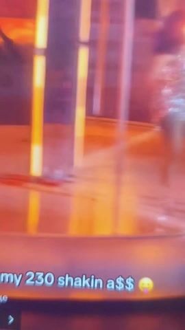 A woman with purple hair and a sparkly outfit dances around a pole. The background is blurry with orange and white lights. The video has text that says "my 230 shakin a$$".