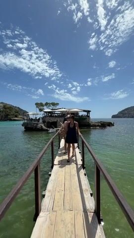 A video shows a wooden pier extending over clear green water towards a small island. On the pier, three people are walking towards the camera. The person in the foreground, a young woman wearing a dark blue dress, sunglasses, and a necklace, is smiling. Behind her, another woman in a white patterned top and sunglasses is also walking. A third person, partially obscured, follows them. On the island, there is a building with an outdoor seating area, shaded by umbrellas and trees. The sky is blue with scattered white clouds, and the water is calm and slightly choppy. Hills covered in vegetation are visible in the background on either side of the island.