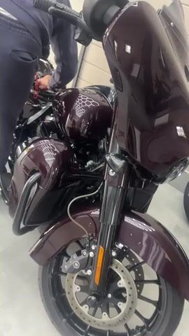 The video shows a close-up, panning shot of a dark red motorcycle. The motorcycle is parked indoors, likely in a showroom, as indicated by the clean, light-colored floor and a light-colored wall in the background. A person's legs, dressed in dark pants, are visible in the upper left corner of the frame, suggesting they are interacting with the motorcycle, possibly preparing to ride it or making adjustments. The motorcycle features a prominent fairing, chrome accents, and a detailed paint job with a hexagonal pattern. The front wheel and brake system are clearly visible. The camera movement is smooth, allowing a good view of the motorcycle's details.