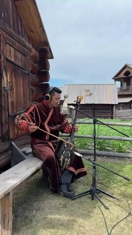 A man in traditional Mongolian attire, a dark maroon velvet robe with intricate gold embroidery, is seated on a wooden bench outdoors. He is wearing headphones and playing a stringed instrument, likely a Morin Khuur, a horsehead fiddle. The instrument has a distinctive carved horsehead at the top. He is also singing, with his mouth open.

The scene appears to be a historical reenactment or cultural demonstration in Shushenskoye, Russia, given the background. Behind him are rustic wooden buildings, possibly log cabins, and a wooden fence. The architecture suggests a rural or historical setting. The weather appears to be overcast, with a cloudy sky, indicating it is likely daytime, though not necessarily bright sunlight. A microphone is set up on a stand in front of the man, suggesting the performance is being recorded. The overall atmosphere seems to be one of cultural expression and performance. There is no visible text in the frame.