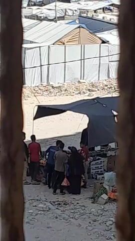 A group of people are gathered under a black tarp. Some are wearing casual clothing, and one person is wearing a dark head covering. They appear to be interacting and possibly exchanging items. The environment looks like a temporary settlement, with white tents and debris in the background. The ground is sandy or dusty.