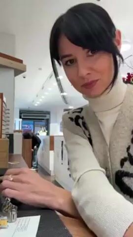 A woman with dark hair pulled back into a ponytail and wearing a white turtleneck and a beige cardigan with black animal print is shown in a mid-shot. She is standing behind a counter in what appears to be an optical shop. She is working on something using a small hammer. The shop is well-lit, and other individuals can be seen in the background. She glances at the camera several times while working.