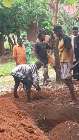 The video shows several people digging a grave in a grassy area with trees. Most of the people are standing around observing while three men are actively digging with shovels. The soil is a reddish-brown color. Some of the people are dressed casually, and some are wearing more formal attire. Most are barefoot or wearing sandals. The background shows trees and possibly houses in the distance.