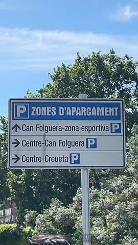 The video opens with a close-up of a blue and white road sign, likely for directing drivers to parking facilities. The top of the sign reads "P ZONES D'APARCAMENT" (Parking Zones), with a large 'P' symbol in blue on a white background. Below this, three parking options are listed with arrows and a 'P' symbol at the end of each: an upward arrow points to "Can Folguera-zona esportiva P" (Can Folguera-sports zone P), while two right-facing arrows indicate "Centre-Can Folguera P" (Center-Can Folguera P) and "Centre-Creueta P" (Center-Creueta P). The sign is positioned against a backdrop of lush green trees and a bright blue sky with scattered white clouds, suggesting a clear day.

The camera then pans slowly to the right, revealing a rural landscape. A narrow stream or canal, reflecting the clear sky, flows through a verdant area filled with tall grasses, reeds, and various trees and bushes. In the middle ground, a large metal electricity pylon stands prominently, with power lines stretching across the sky. More green foliage and distant, hazy hills or mountains complete the background. The overall impression is one of a natural environment interspersed with elements of infrastructure. The weather is sunny and bright, indicating daytime. The scene is located in Santa Perpètua de Mogoda, Spain.