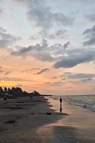 The video shows a beach at sunset. The sky is a mix of orange, pink, and grey with a crescent moon visible. The beach is dark sand, and the water is coming in gently. There are a few people walking along the beach, and one person is standing at the edge of the water, their reflection visible in the wet sand. In the background, there are trees and possibly some buildings. The sound of a motorcycle can be heard.