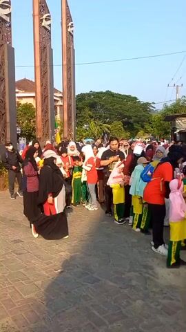 A group of people, mostly women and children, are gathered outdoors on a paved walkway. The children are wearing uniforms that consist of green pants and yellow shirts, with some wearing white headscarves. Many of the adults are also wearing headscarves. The group appears to be in a line or a loosely organized crowd. In the background, there are trees, buildings, utility poles, and what appear to be large decorative pillars. The sky is clear and blue, suggesting it is daytime and likely sunny. Some of the people are holding small flags. The overall impression is that of an organized event or outing.