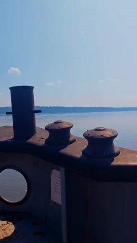 The video pans across the deck of a boat on a sunny day. The camera focuses on a railing with several large metal bollards, used for tying up ropes. Two cylindrical bollards stand prominently on the left, followed by two winch-like devices with spools. In the foreground, a circular window is visible on the side of the boat, reflecting the sky and water. The background shows a calm body of water stretching to a distant tree-lined shore under a clear blue sky with scattered clouds. A small part of a mechanical arm or winch assembly is visible in the lower left corner. The overall impression is one of a leisurely voyage or a stationary boat on a bright, clear day.