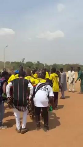 The video shows a group of people dancing in a circle on a sunny day. Most of the dancers are wearing yellow shirts with black writing on them. Some are dressed in traditional clothing, with fringed garments. Some are holding hands and dancing together, while others are moving individually. Several people standing outside the circle are recording the dance with their phones. The environment is a dirt ground with some trees in the background.