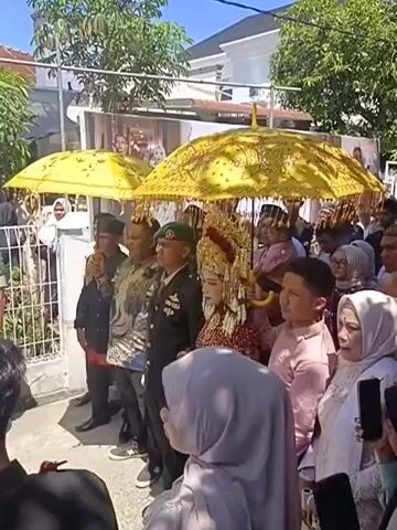 A wedding procession is captured in this video. A bride, adorned in traditional wedding attire and a tall, ornate headdress, stands prominently. She is accompanied by a military officer in uniform and other individuals, some holding large, decorative yellow umbrellas to shield the bride. A crowd of onlookers, many of whom are women wearing hijabs, observes the procession. In the background, a white house and trees are visible, suggesting an outdoor setting. The overall atmosphere appears festive, with people gathered to celebrate the wedding.
