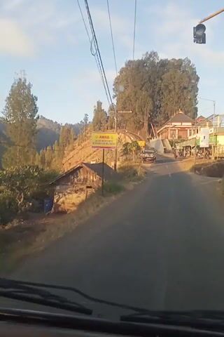 The video shows a road view from inside a vehicle. A sign with the text "BRAMO HILLSIDE" is visible. There are houses, trees, and utility poles along the road. Some people are seen near the houses. A motorcycle is parked near a building with shops. The road appears to be in a rural or semi-rural area with hills in the background. The sun is shining, indicating it is daytime. The camera angle is tilted, making the scene appear sideways.