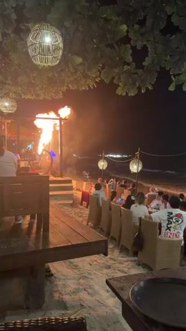 This video captures a beachside restaurant at night. In the foreground, a wooden table is visible on the right, and a wooden bench with a person seated on it is on the left. Overhead, woven spherical lanterns hang from trees. In the mid-ground, a performance space features a large metal sphere with flames emanating from it, creating a fiery spectacle. Several people are gathered around this area. To the right, numerous patrons are seated at tables on the sandy beach, facing the ocean. The ocean waves are visible in the background, and lights from a distant area on the water can be seen. The overall atmosphere is dark, illuminated by the fire and various artificial lights.