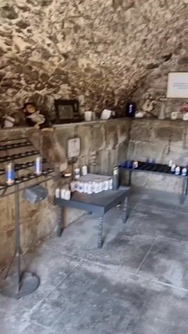The video pans across a small, dimly lit stone room that appears to be a chapel or shrine. The walls are rough-hewn stone, and the floor is made of large stone tiles. Along the back wall, a shelf holds various religious items including candles, crosses, flowers, and framed pictures. There are also two dark tables in front of the shelf, each with rows of votive candles. To the right, there is a wooden lectern and a wooden chair. The overall impression is one of solemnity and quiet devotion.