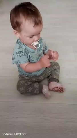 A toddler sits on a light-colored wooden floor with a pacifier in their mouth. The toddler is wearing a light blue t-shirt with a pattern of toy trucks and green pants with a similar pattern. They have pink socks on their feet with red polka-dot grips on the soles. The toddler is holding a clear object, possibly a toy, in their hands and appears to be examining it. The background is mostly the floor with some indistinct shadows. The camera is positioned slightly above and to the side of the toddler.