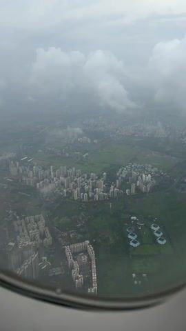 The video shows an aerial view of a densely populated urban area with numerous buildings and green spaces, likely taken from an airplane window. The sky is overcast with clouds, suggesting a hazy or rainy day. In the foreground, a portion of the airplane's circular window frame is visible, indicating the perspective. The landscape below is a mix of residential buildings, some appearing to be apartment complexes, and large expanses of green land, possibly parks or undeveloped areas. In the middle ground, a cluster of tall buildings forms a distinct urban center. Further in the distance, the urban sprawl continues, becoming more diffused and less distinct due to the atmospheric conditions and distance. There are no people or specific actions clearly visible from this altitude and through the haze. The overall impression is of a sprawling city seen from above on a cloudy day.