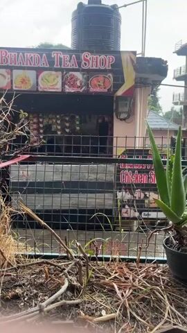 The video opens with a view of a roadside food stall labeled "Bhakda Tea Shop" in Fatehpur, India. The stall appears to sell a variety of food items, including tea, omelets, and chicken dishes, as indicated by the signage. Behind the stall, a large black water tank is visible.

The camera then shifts to a close-up of a weathered wooden table. On the table, several objects are laid out: a smartphone, a small black packet labeled "FAIRLAND," a small ashtray with some sticks, a white cloth-wrapped item, a rectangular beige tray, and a small white bowl.

Further shots reveal a plate with sushi rolls and chopsticks, a small dipping bowl of sauce, and a glass of water. Another shot shows a red bowl filled with noodles and a half-boiled egg, with a spoon resting in it. Finally, a red plate with noodles and an egg is shown, along with a small glass of red sauce. The scene suggests a casual meal being enjoyed outdoors, possibly at a roadside eatery.

The lighting suggests it is daytime, with overcast skies. The presence of an aloe vera plant in a pot in the foreground adds a touch of greenery to the scene. The overall atmosphere appears relaxed and informal. The food items, including sushi and noodles, indicate a diverse culinary offering.