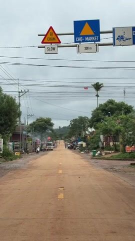 The video shows a view down a long, straight, dirt road. There are buildings and trees on either side of the road, and some people and vehicles are visible in the distance. There are traffic signs hanging over the road. The sky is cloudy.