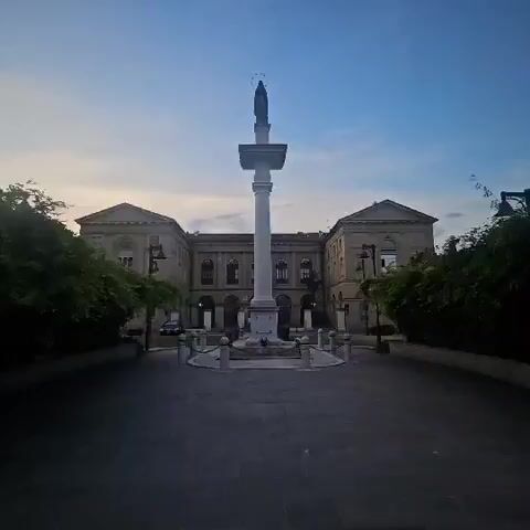 A statue of a woman with a halo stands atop a column in front of a building with trees on either side. The sky is a pale blue. The statue is made of stone and the column is white. The building appears to be made of stone and has arched windows. The trees are green and leafy. The ground in front of the column is paved with dark stones. The statue is a religious monument.