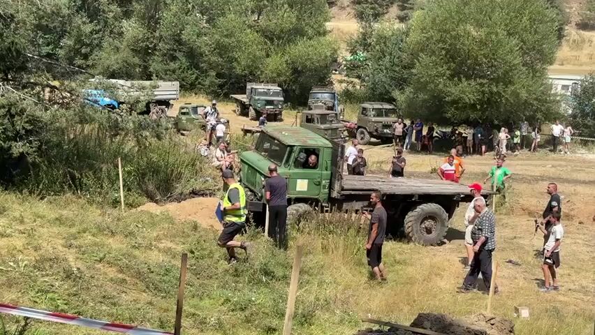 A group of people are gathered around a green flatbed truck that is stuck in a ditch during an off-road event. The truck's front wheels are submerged in mud and dirt, and several people are attempting to free it. Some are pushing on the back of the truck, while others are directing the driver. The truck appears to be an older model, possibly military surplus. In the background, there are other trucks and vehicles, as well as more spectators. The environment is a grassy, wooded area with a dirt track. The weather is sunny and warm.