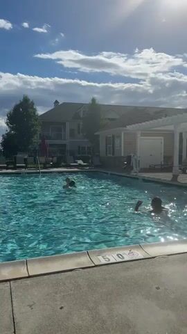 Two people are playing with a ball in a swimming pool on a sunny day. The pool is surrounded by a concrete deck and lounge chairs. In the background, there is a large building with a balcony and trees. The sky is blue with scattered clouds. The person on the left is throwing the ball to the person on the right, who is reaching out to catch it.