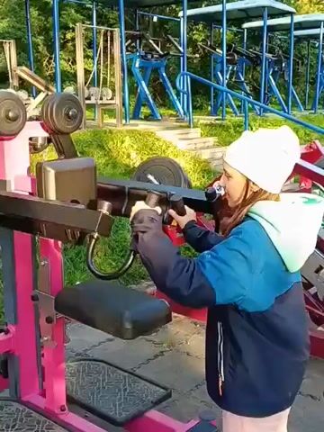 Two young girls are at an outdoor gym. The girl in the foreground, wearing a white beanie, blue and green jacket, and pink pants, is standing in front of a pink weight machine. She is holding onto the machine's handles. The second girl, wearing a brown jacket, stands behind the first girl and has her hands on the first girl's shoulders and back, appearing to assist her. The gym is equipped with multiple blue workout machines lined up in the background. The ground is covered with a dark rubber matting. The environment is outdoors, with trees and foliage visible behind the gym equipment. The lighting suggests it is daytime, possibly late afternoon due to the golden hue.