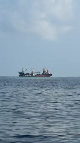 The video depicts a cargo ship at sea. The ship is black, white, and red. It has several cranes or booms extending from its deck. The sea is dark blue and choppy, and the sky is a light blue with some scattered clouds. The ship is moving across the frame from left to right. The horizon line is visible behind the ship.